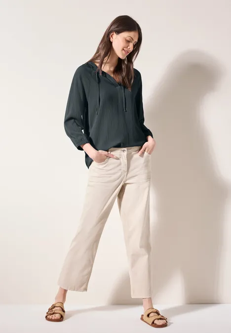 Frau mit dunkler Bluse, heller Hose und Sandalen steht lässig an einer Wand.